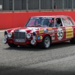 The 1971 AMG 300 SEL 6.8 "Red Pig" in a vintage racing setting, showcasing its boxy sedan shape and red paint.