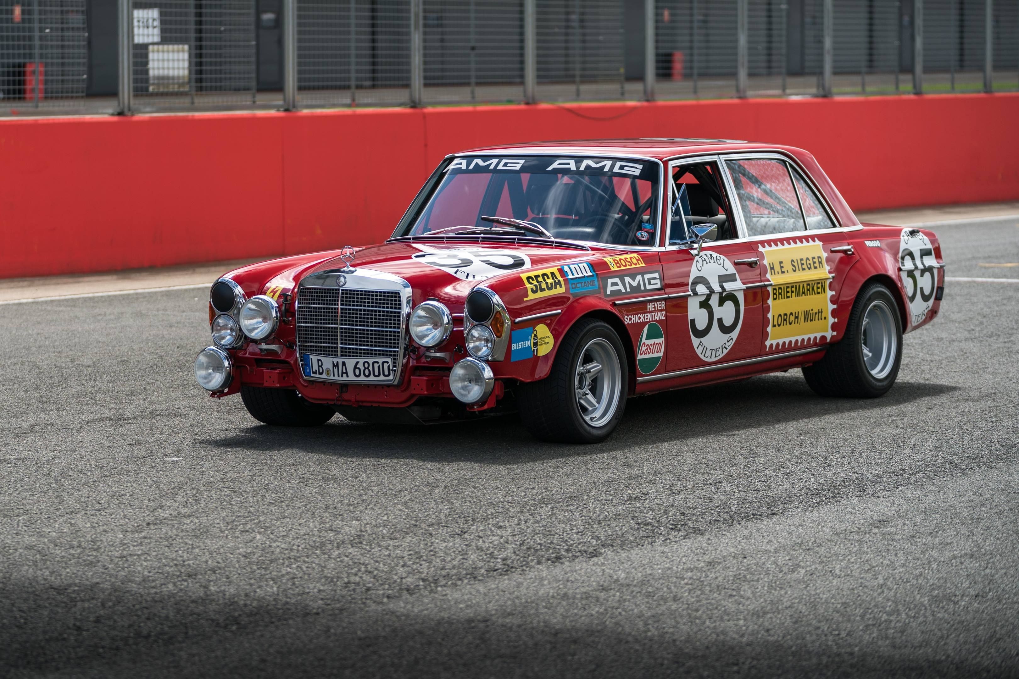 The 1971 AMG 300 SEL 6.8 "Red Pig" in a vintage racing setting, showcasing its boxy sedan shape and red paint.